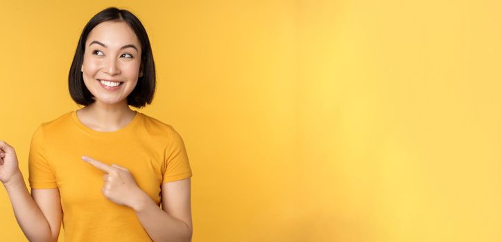 Portrait of happy asian girl pointing fingers and looking left, smiling amazed, checking out promo banner, showing advertisement against yellow background.