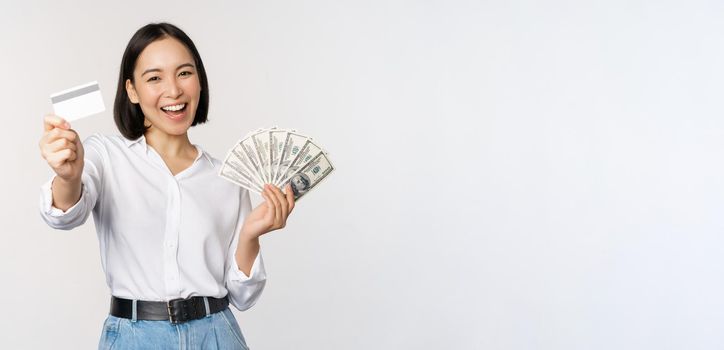 Happy korean woman holding credit card and money dollars, smiling and laughing, posing against white studio background.