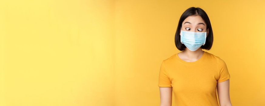 Health and people concept. Funny asian girl squinting, looking at her face medical mask, standing over yellow background.
