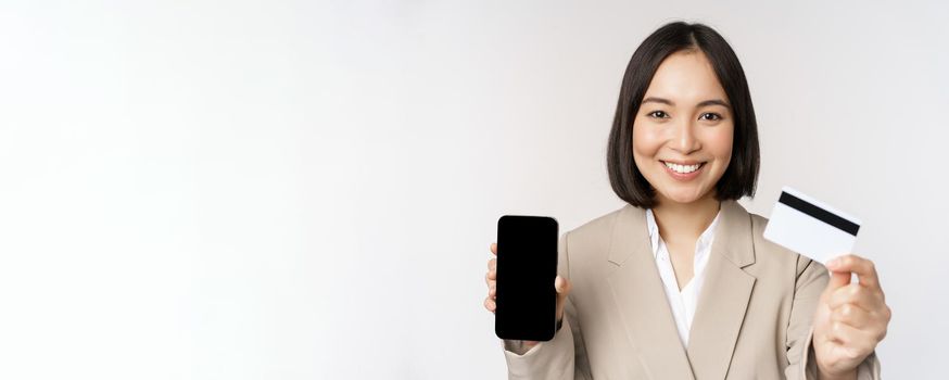 Smiling corporate woman in suit, showing mobile phone screen and app on mobile phone, smartphone screen, standing over white background.