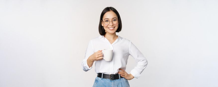 Happy young energetic asian woman smiling, drinking, holding cup mug of coffee, standing confident against white background.