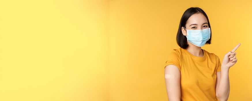 Vaccination from covid and health concept. Image of smiling korean girl in medical face mask, band aid on shoulder, pointing finger at banner advertisement, yellow background.