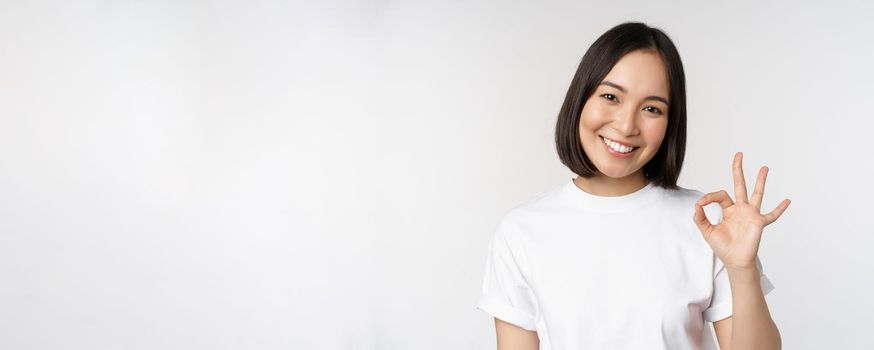 Everything okay. Smiling young asian woman assuring, showing ok sign with satisfied face, standing over white background. Copy space