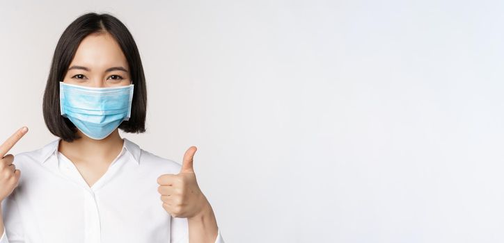 Portrait of asian girl in medical mask showing thumbs up sign and pointing at her covid protection, standing over white background.