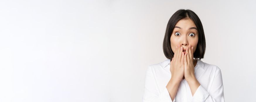 Close up face of asian woman gasping, looking shocked and speechless, holding hands near mouth and staring at camera impressed, white background.