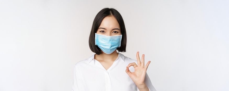 Covid and health concept. Portrait of asian woman wearing medical face mask and showing okay sign, standing over white background.