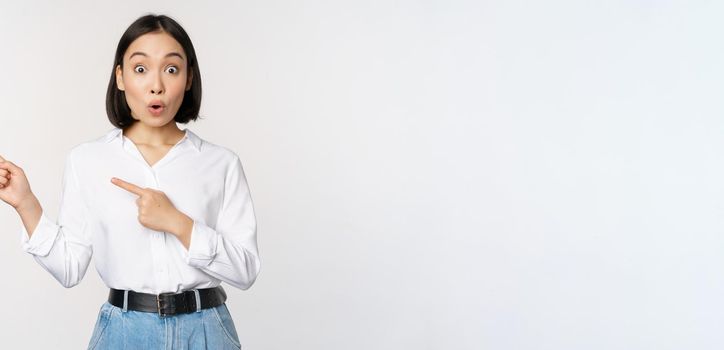 Portrait of excited young asian woman, office lady pointing fingers left at discount, showing sale banner, standing over white background.