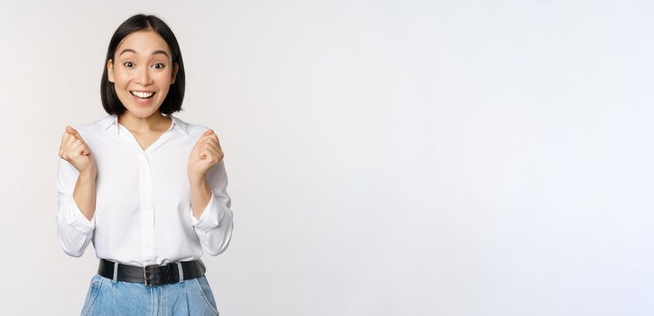 Enthusiastic asian woman rejoicing, say yes, looking happy and celebrating victory, champion dance, fist pump gesture, standing over white background.