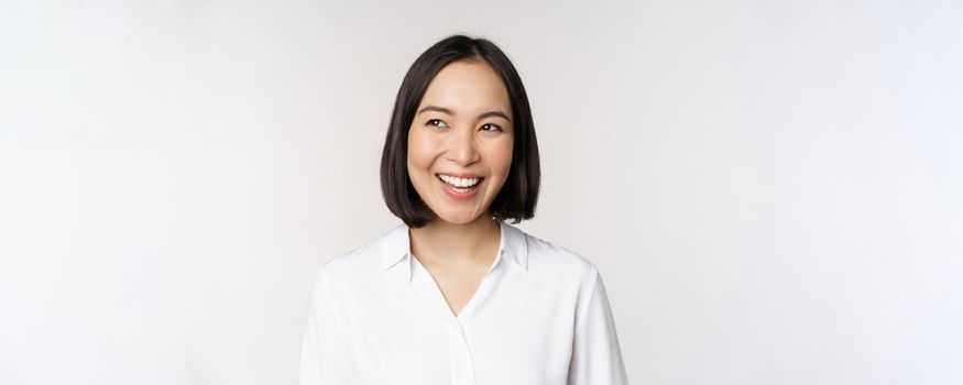 Image of smiling asian woman planning, thinking of smth, daydreaming, standing over white background with smug face. Copy space