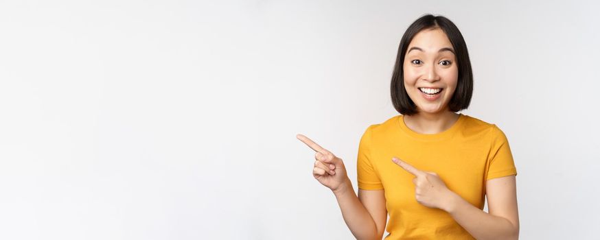 Portrait of smiling asian brunette girl in yellow tshirt, pointing fingers left, showing copy space, promo deal, demonstrating banner, standing over white background.