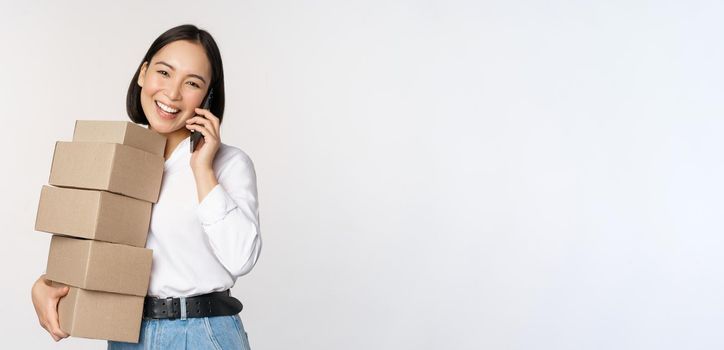 Image of young asian businesswoman answer phone call while carrying boxes for delivery, posing against white background.