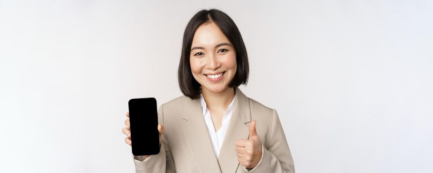 Smiling asian woman showing smartphone screen and thumbs up. Corporate person demonstrates mobile phone app interface, standing over white background.