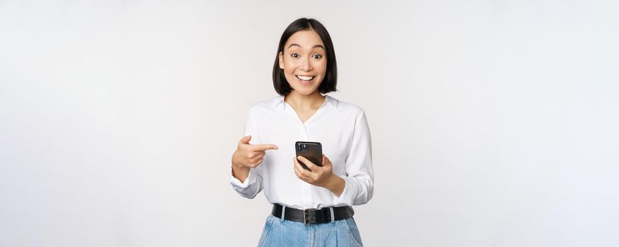 Amazed girl talking about smartphone app, pointing at phone while looking impressed at camera, standing against white background.