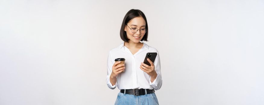 Image of modern asian woman looking at mobile phone, drinking takeaway coffee, wearing glasses, using smartphone app, standing over white background.