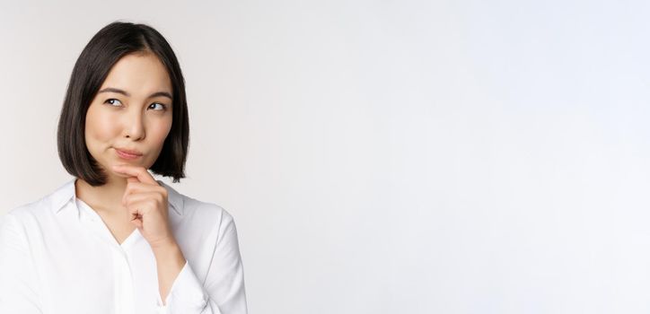 Close up portrait of asian woman thinking, looking aside and pondering, making decision, standing over white background.
