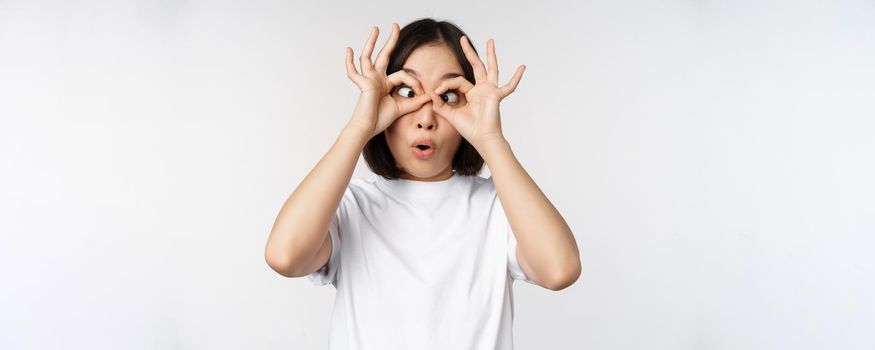 Funny young asian woman, korean girl making eyes glasses gesture, looking happy at camera, standing over white background.