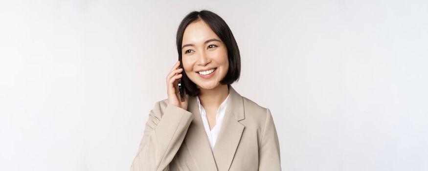Smiling corporate woman in suit, talking on mobile phone, having a business call on smartphone, standing over white background.