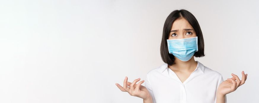 Distressed and miserable young asian woman in face mask, looking up, looking up sad, standing over white background.