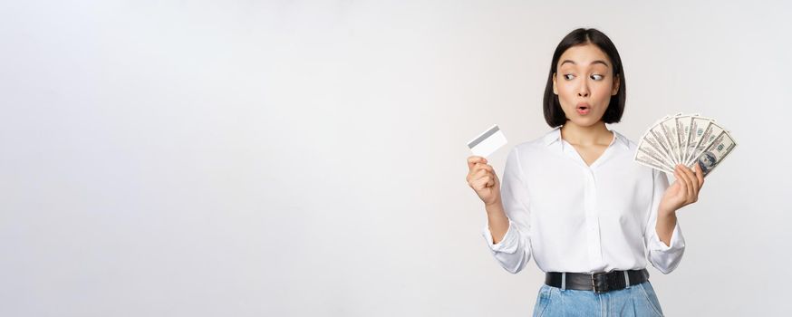Excited korean girl looking at credit card, holding money cash, posing against studio background. Finance concept
