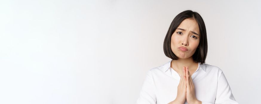 Close up portrait of asian woman begging, asking for smth, say please with coy sad face, need help, standing over white background.