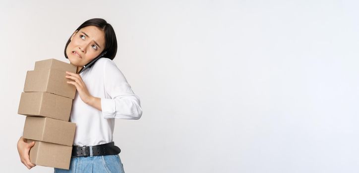 Young asian businesswoman answer phone call, talking on mobile while carrying pile of boxes with orders, standing over white background.