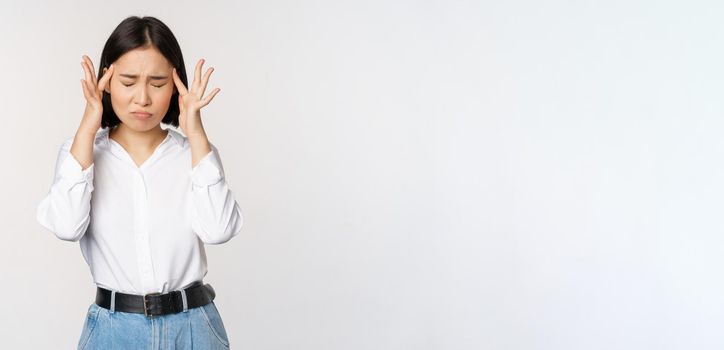 Image of distressed young asian businesswoman, female lady touching head, rubbing temples, has migraine, standing over white background.