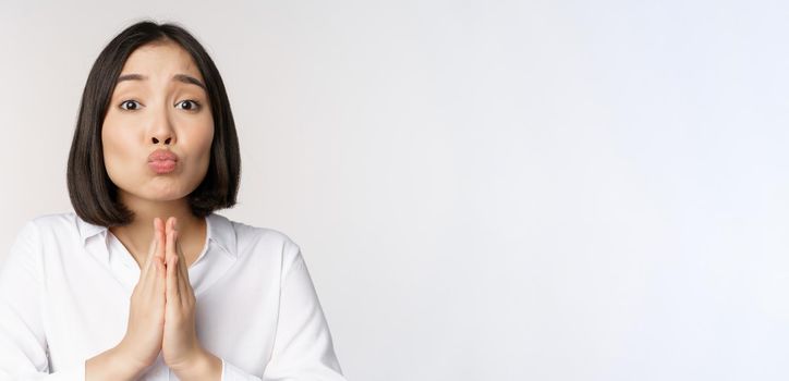 Cute asian woman begging, say please, asking for favour, need help, standing with coy face against white background.