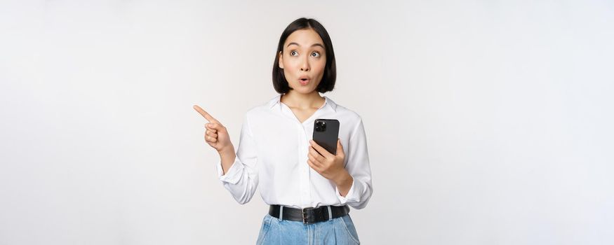 Excited asian girl shopping on mobile app, holding phone and pointing finger left at empty copy space, showing promo text, standing with smartphone against white background.