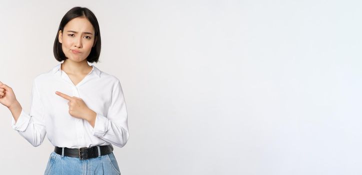 Portrait of skeptical asian woman frowning, pointing fingers left and complaining, express dislike or disapproval, standing against white background.