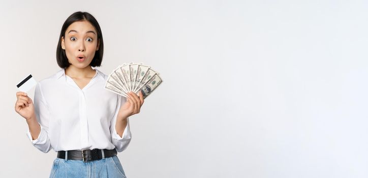 Finance and money concept. Happy young asian woman dancing with cash and credit card, smiling pleased, posing against white studio background.