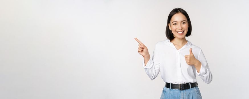 Image of young asian business woman, smiling while pointing finger left and showing thumbs up, recommending product, praise, standing over white background.