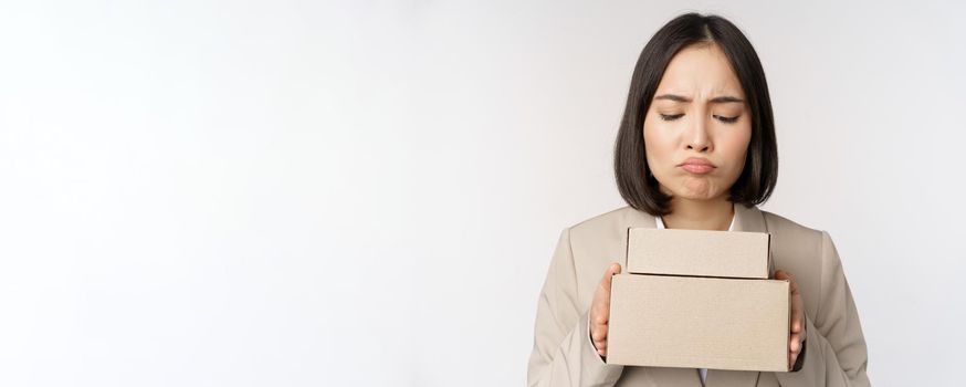 Portrait of asian saleswoman, female entrepreneur holding boxes and looking sad, disappointed, standing over white background.