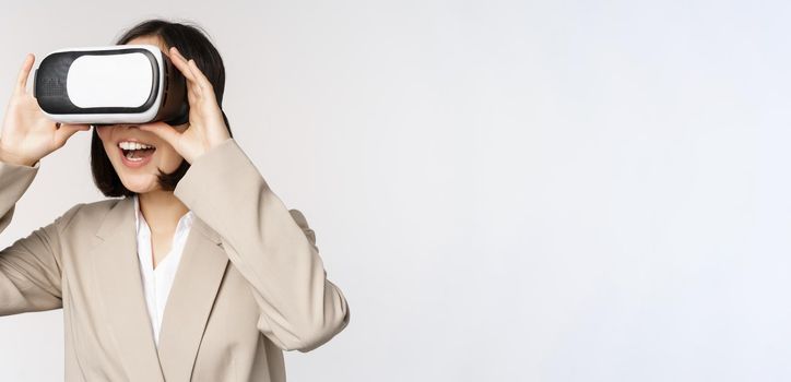 Amazed business woman in suit using virtual reality glasses, looking amazed in vr headset, standing over white background.