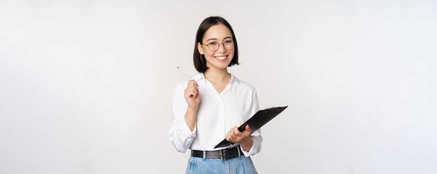 Smiling young asian woman taking notes with pen on clipboard, looking happy, standing against white background.