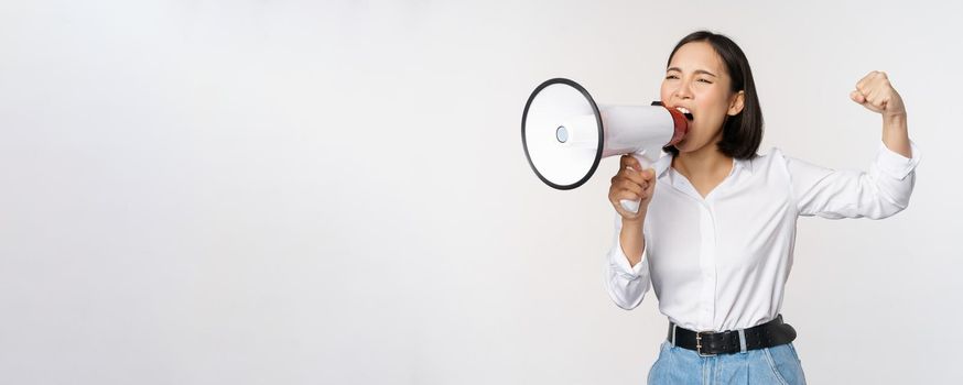 Enthusiastic asian woman, girl activist shouting at protest, using megaphone, looking confident, talking in loudspeaker, protesting, standing over white background.