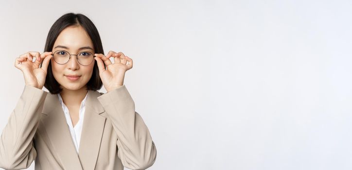 Smiling asian businesswoman trying new glasses, wearing eyewear, standing in suit over white background. Copy space