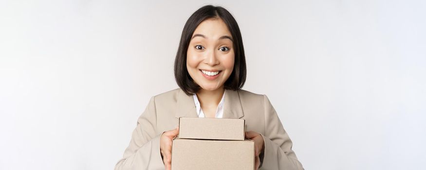 Image of asian saleswoman, business woman giving boxes with order, deliver to customer, standing in suit over white background.