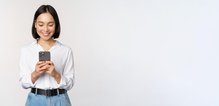 Stylish modern asian girl using mobile phone application, chatting on cellphone and smiling, standing in white blouse against studio background.