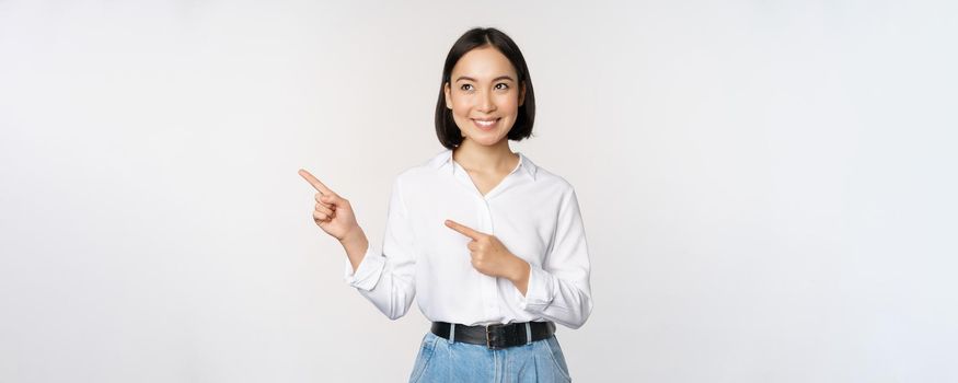 Portrait of attractive adult asian woman pointing, looking left with pleased smile, showing banner or logo aside, standing against white studio background.