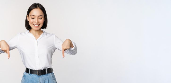 Portrait of happy asian woman pointing fingers down and looking below at advertisement, showing info banner, advertising, standing over white background.