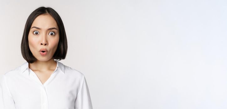 Image of korean woman looking surprised and happy at camera, standing over white background. Copy space