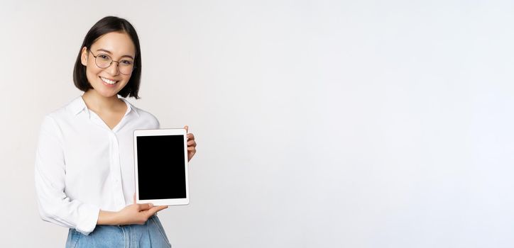 Young asian woman professional demonstrates digital tablet screen, info on her gadget, smiling and looking at camera, standing over white background.