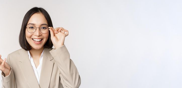 Enthusiastic asian saleswoman in glasses, smiling and laughing, looking amazed at camera, standing in beige suit over white background.