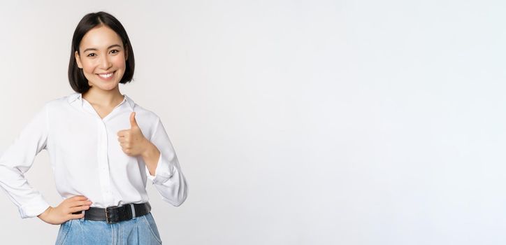 Image of confident asian woman showing thumb up in approval, recommending, like smth good, standing over white background.