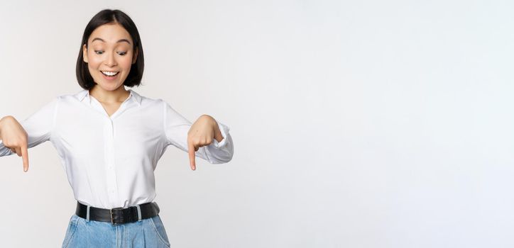 Portrait of happy asian woman pointing fingers down and looking below at advertisement, showing info banner, advertising, standing over white background.