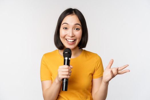 Image of young asian woman talking in microphone, perfom with mic, giving speech, standing in yellow tshirt against white background.