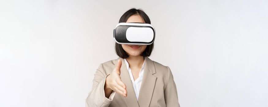 Meeting in vr chat. Asian businesswoman in virtual reality glasses, extending hand for handhshake with business partner, greeting someone, standing over white background.