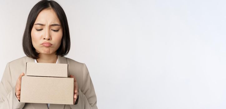 Portrait of asian saleswoman, female entrepreneur holding boxes and looking sad, disappointed, standing over white background.