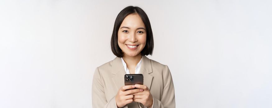 Image of asian businesswoman in suit, holding mobile phone, using smartphone app, smiling at camera, white background.