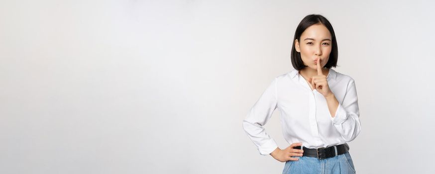 Portrait of young asian adult woman shushing, say hush shh, press finger to lips, sharing secret, dont speak taboo gesture, standing over white background.